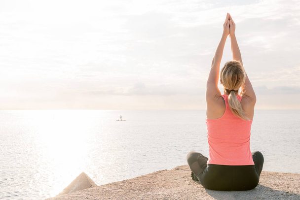 back view of an unrecognizable blonde woman practicing yoga at sunrise sitting in front of the sea, concept of mental health and relaxation, copy space for text - Photo, image