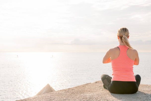 back view of an unrecognizable blonde woman doing yoga at dawn sitting in front of the sea, mental health care and relaxation concept, copy space for text - Photo, Image