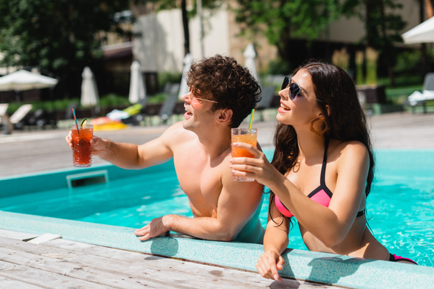 vrolijk paar in zonnebril holding cocktails in zwembad  - Foto, afbeelding