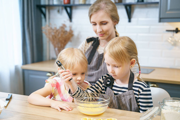 Two cute sisters helping mom in the kitchen. Happy family preparing dough for cookies. - Photo, Image