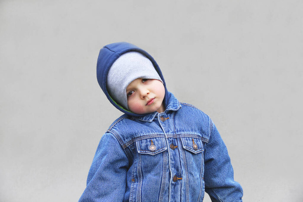 A boy in a jacket and hat is looking at the camera. Fashionable child on a gray background. The guy in the denim jacket. - Фото, изображение