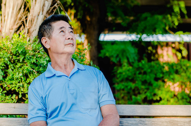Asiatischer älterer Herr denkt nach, während er aufblickt und auf einer Holzbank sitzt und es sich an einem sonnigen Tag in einem Park gemütlich macht - Foto, Bild