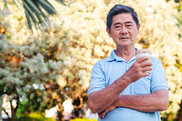 Azjatycki starszy pan stoi w parku i pije kawę z relaksującym się w słoneczny dzień - Zdjęcie, obraz