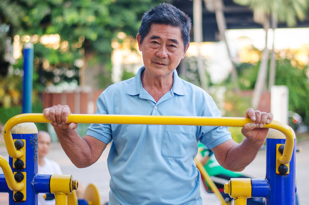 Asian senior gentleman making exercises on outdoor gym against green summer park as a background - Photo, Image