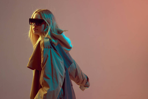 Stylish young girl, cool posing in a hoodie, sunglasses and cloak with developing hair, on a neon background. - Foto, imagen