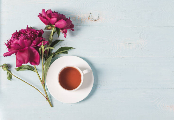 Κόκκινο ροζ παιώνιες φλιτζάνι τσάι σε μπλε ξύλινο φόντο. Οριζόντιος χώρος αντιγραφής πλαισίου για σχεδιασμό κειμένου. Πάνω όψη επίπεδη διάταξη - Φωτογραφία, εικόνα