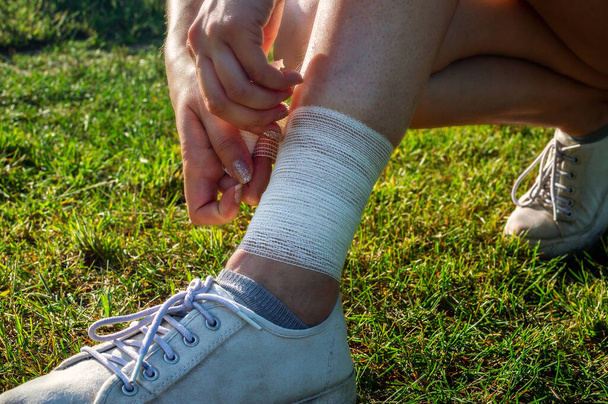 Young woman bandaging injured ankle. Injury leg while running outdoors. First aid for sprained ligament or tendon - Photo, Image