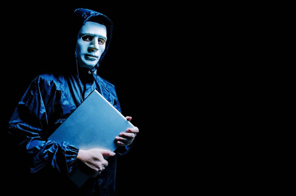 Anonimowy haker komputerowy w białej masce i bluzie. Obscured ciemna twarz trzyma laptopa mocno w rękach, Złodziej danych, atak internetowy, dark net i cyberbezpieczeństwa koncepcji. - Zdjęcie, obraz