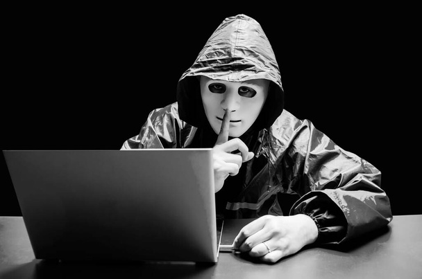 Anonymer Computerhacker in weißer Maske und Kapuzenpulli. Verdunkelte dunkle Gesicht machen Schweigegeste versuchen zu hacken und zu stehlen Informationssystem Daten von Computer mit USB-Stick auf dunklem Hintergrund, Datendieb, Internet-Angriff. - Foto, Bild