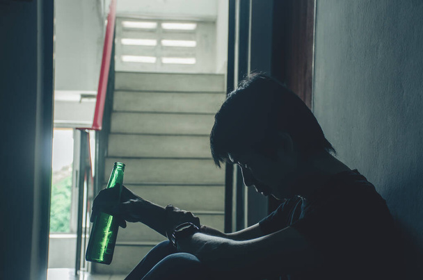 Het silhouet van jonge mannen met een fles alcoholbier in de oude flat, Ongezonde Lifestyle, Verslaafde mannen, Alleen zitten, Depressieve man met fles, Verslaafd aan alcohol. - Foto, afbeelding