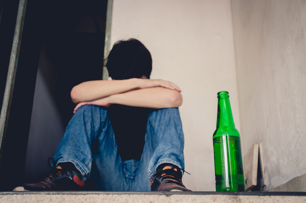 Nieszczęśliwy samotny młody człowiek pijący piwo i alkohol czuje się zmarnowany, samotny i zdesperowany w bólu i stresie emocjonalnym, nałogu i męskiej depresji. Selektywne skupienie się na butelce piwa - Zdjęcie, obraz