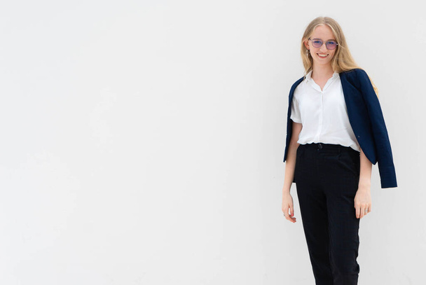 A portrait of young caucasian blonde business woman in glasses standing in front of a white wall, smiling and looking to the camera. Her hands are down. She is dressed in black pants, blue jacket and a white short-sleeved shirt. Concept of successful - Foto, afbeelding