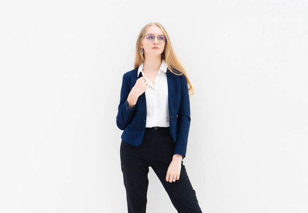 A portrait of young caucasian blonde business woman in glasses standing in front of a white wall and looking away. The right hand holds a jacket. She is dressed in black pants, blue jacket and a white short-sleeved shirt. Concept of successful young  - Фото, изображение