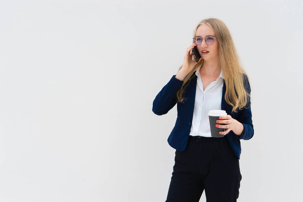 Portrait of a young European blonde business woman is standing in front of a white wall, smiling, talking to the phone and holding coffee in her hand. She is dressed in black pants, blue jacket and a white short-sleeved shirt. Surprised facial expres - Foto, imagen
