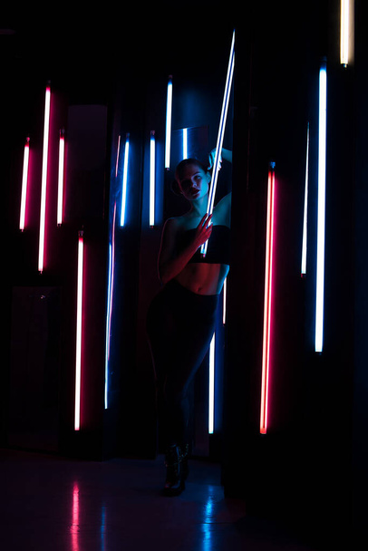Full body sensuele vrouw met heldere lamp leunend op de muur in de donkere kamer met neon verlichting - Foto, afbeelding