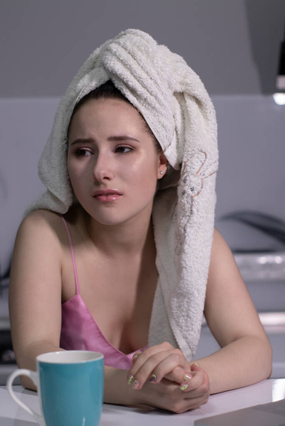 triest, aantrekkelijk brunette meisje - in roze pyjama en met een witte handdoek op haar hoofd - bijna huilen- in een grijze keuken. - Foto, afbeelding