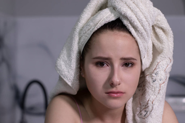 λυπημένη, ελκυστική μελαχρινή κοπέλα - με ροζ πιτζάμες και με λευκή πετσέτα στο κεφάλι - σχεδόν κλαίγοντας - σε μια γκρίζα κουζίνα. - Φωτογραφία, εικόνα