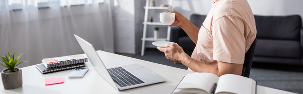 Panoramic shot of man holding cup and saucer near laptop and stationery on table at home, earning online concept - Foto, imagen
