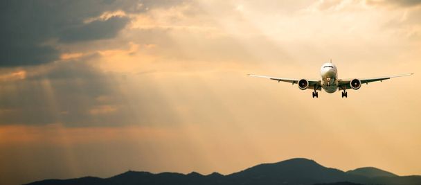 Επιβατικό αεροπλάνο. Τοπίο με το μπροστινό μέρος του λευκού αεροπλάνου πετά στον πορτοκαλί ουρανό με σύννεφα πάνω από τα βουνά, θάλασσα στο πολύχρωμο ηλιοβασίλεμα. Τα επιβατικά αεροσκάφη προσγειώνονται. Εμπορικό αεροπλάνο. Ιδιωτικό τζετ. Ταξίδι - Φωτογραφία, εικόνα