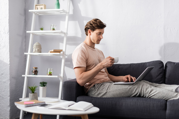 Selektywna ostrość przystojnego freelancera trzymającego filiżankę kawy i korzystającego z laptopa w pobliżu notebooków na stoliku kawowym, zarabiającego koncepcję online - Zdjęcie, obraz
