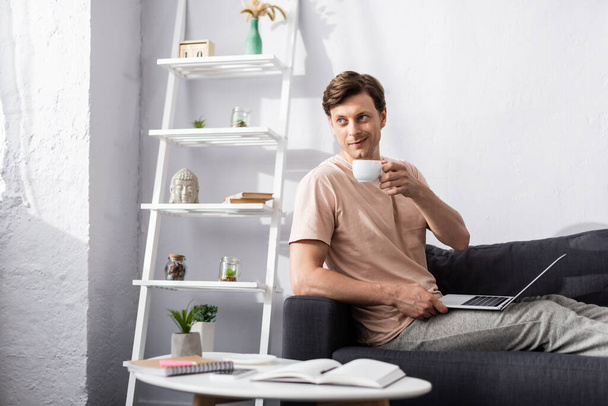 Foco seletivo de sorrindo freelancer beber café e usando laptop perto de artigos de papelaria na mesa de café, ganhando conceito online
 - Foto, Imagem