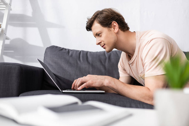Selective focus of handsome freelancer using laptop on couch at home, earning online concept - 写真・画像