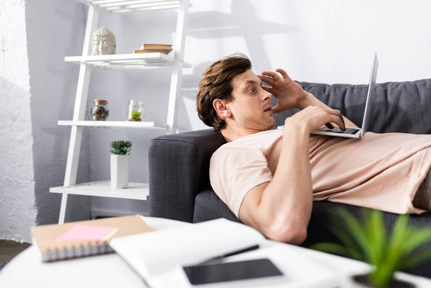 Вибірковий фокус працівника з використанням ноутбука, лежачи на дивані біля блокнотів і смартфона на кавовому столі, заробляючи онлайн концепцію
 - Фото, зображення