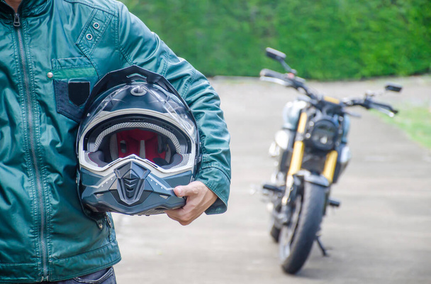 Vorderansicht des gutaussehenden Fahrers Biker Mann in grüner Lederjacke hält einen Helm und Vintage-Motorrad verschwimmen Hintergrund - Foto, Bild