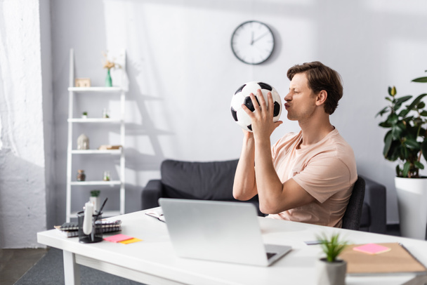 Valikoiva painopiste mies suudella jalkapalloa istuessaan lähellä kannettavaa tietokonetta ja paperitavarat pöydällä kotona, käsite ansaita verkossa - Valokuva, kuva