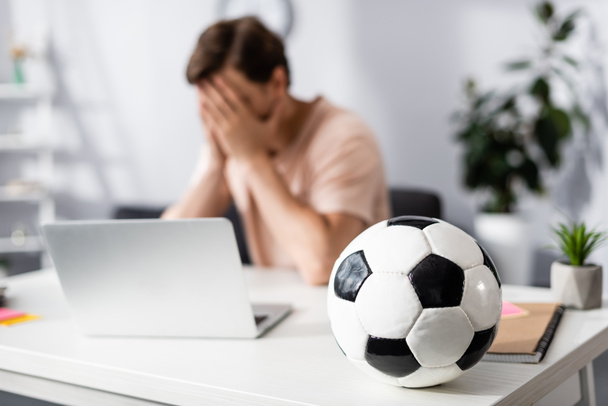 Selective focus of football on table and sad man covering face with hands near laptop at home, earning online concept - Photo, Image