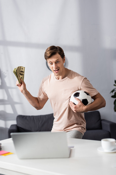 Selectieve focus van opgewonden man met contant geld en voetbal in de buurt laptop op tafel thuis, concept van het verdienen van online - Foto, afbeelding