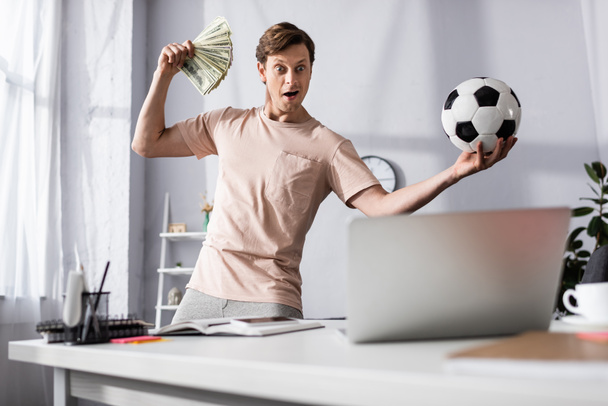 Focus selettivo di uomo scioccato con denaro contante e calcio guardando computer portatile in salotto, concetto di guadagnare online - Foto, immagini