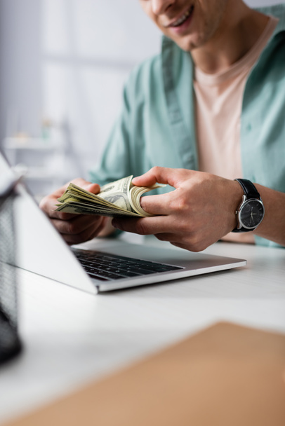 Rajatut näkymä hymyilevä mies laskee dollareita lähellä kannettavaa pöydällä kotona, ansaita online käsite - Valokuva, kuva