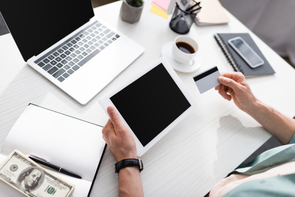 Cropped view of man holding credit card and digital tablet with blank screen near dollars and notebook on table, earning online concept - Foto, imagen