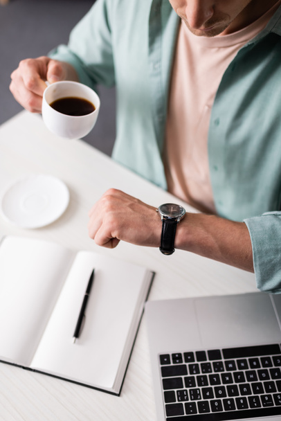 Обрезанный вид фрилансера время проверки на наручные часы во время питья кофе возле ноутбука и ноутбука, концепция управления временем  - Фото, изображение