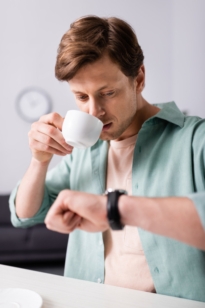 ハンサムな男がコーヒーを飲み、自宅で腕時計の時間をチェックする選択的な焦点、時間管理の概念  - 写真・画像