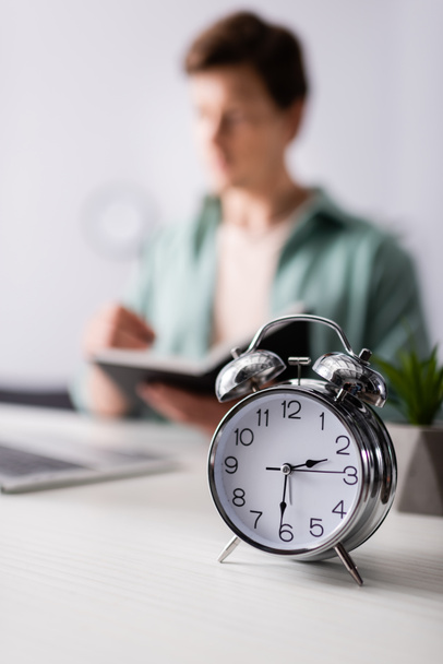 Selective focus of alarm clock on table and man holding notebook near laptop at home, concept of time management  - Foto, imagen