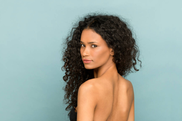 Beauty portret van een Afro-Amerikaanse vrouw met prachtige lange krullend haar draaien om terug te kijken over haar schouder op de camera tegen een blauwe studio achtergrond - Foto, afbeelding