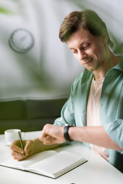 Selectieve focus van glimlachende freelancer controleren van de tijd tijdens het schrijven op notebook in de buurt van kopje koffie, concept van time management  - Foto, afbeelding