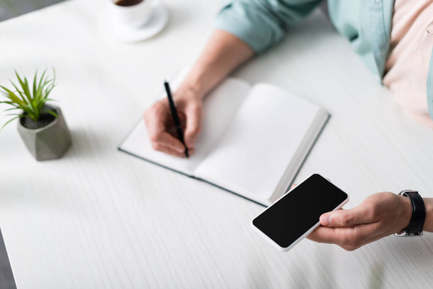 Cropped view of man holding smartphone with blank screen while writing on notebook at table, earning online concept - Photo, Image