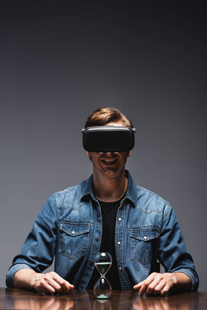 Selective focus of smiling man in vr headset sitting near hourglass on table on grey background, concept of time management  - Fotografie, Obrázek