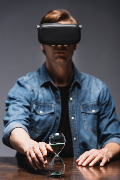 Selective focus of man in virtual reality headset pulling hand to hourglass on table on grey background, concept of time management  - Fotó, kép