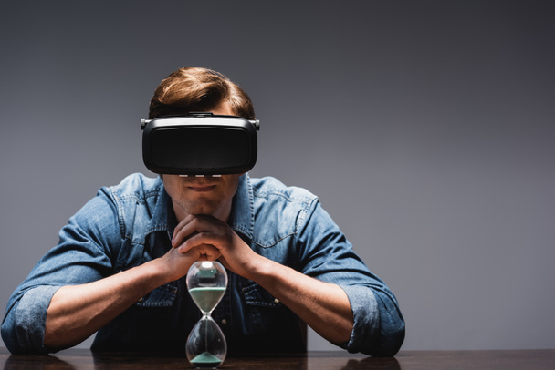 Man in vr headset sitting near hourglass on table on grey background, concept of time management  - Photo, Image