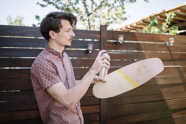Young man spraying paint over wooden skateboard - Fotoğraf, Görsel