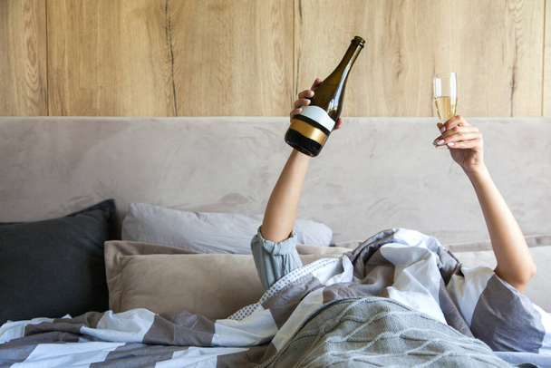 Young woman waking up in the morning in the bed, hidding under the blanket, stretching out one's arms with a cup of coffee and showing peace sign. alcohol, celebration and morning concept - hand of young woman lying in bed with champagne glass - Photo, Image