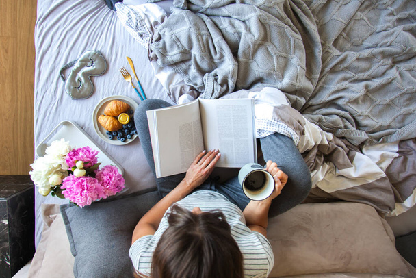 Desayuno con cruasanes y café servido en la cama gris. Hermosa joven está acostada en la cama y leyendo un libro
. - Foto, imagen