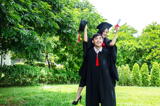 Ένας άντρας και μια γυναίκα ζευγάρι ντυμένοι στα μαύρα φόρεμα αποφοίτησης ή απόφοιτοι με συγχαρητήρια με καπέλα αποφοίτησης στέκεται, αυτή τον αγκαλιάζει από πίσω χαμογελά και κρατώντας το καπέλο με το φόντο πάρκο - Φωτογραφία, εικόνα