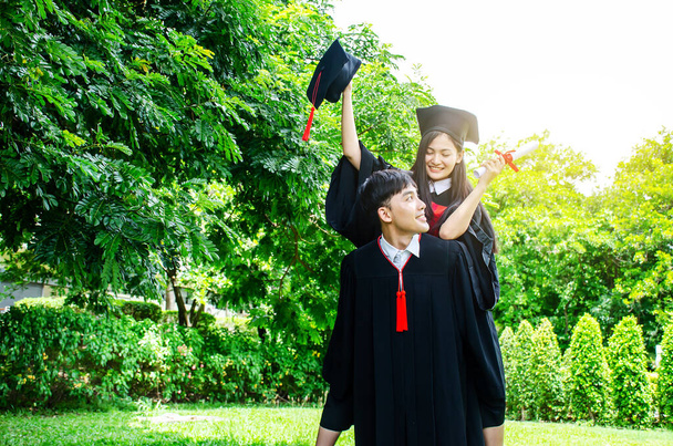 Ein Paar im schwarzen Abschlusskleid oder Absolventen mit Glückwünschen mit Abschlussmützen steht da, sie umarmt ihn von hinten, lächelt und hält den Hut mit dem Park - Foto, Bild