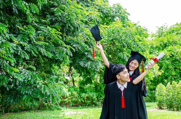 Um homem e mulher casal vestido com vestido de graduação preto ou graduados com parabéns com chapéus de formatura está de pé, ela está abraçando-o pelas costas está sorrindo e segurando chapéu com o parque
 - Foto, Imagem