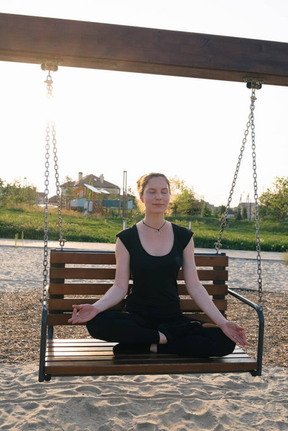 beautiful young woman meditating on a swing in a city park at the sunset - Фото, изображение